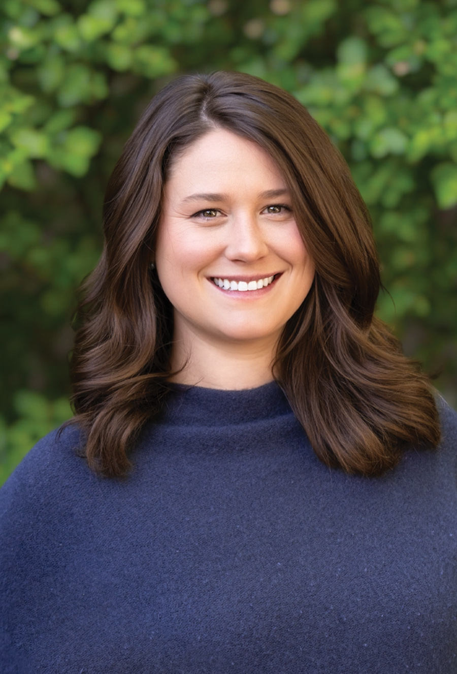 Elizabeth Ferguson, a woman with shoulder-length brown hair and a navy blue top, smiles outdoors in front of green foliage.