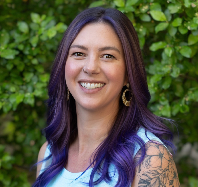 Lorna Bushong, with long purple hair and a floral tattoo on her shoulder, smiles at the camera as she stands in front of green leafy bushes.