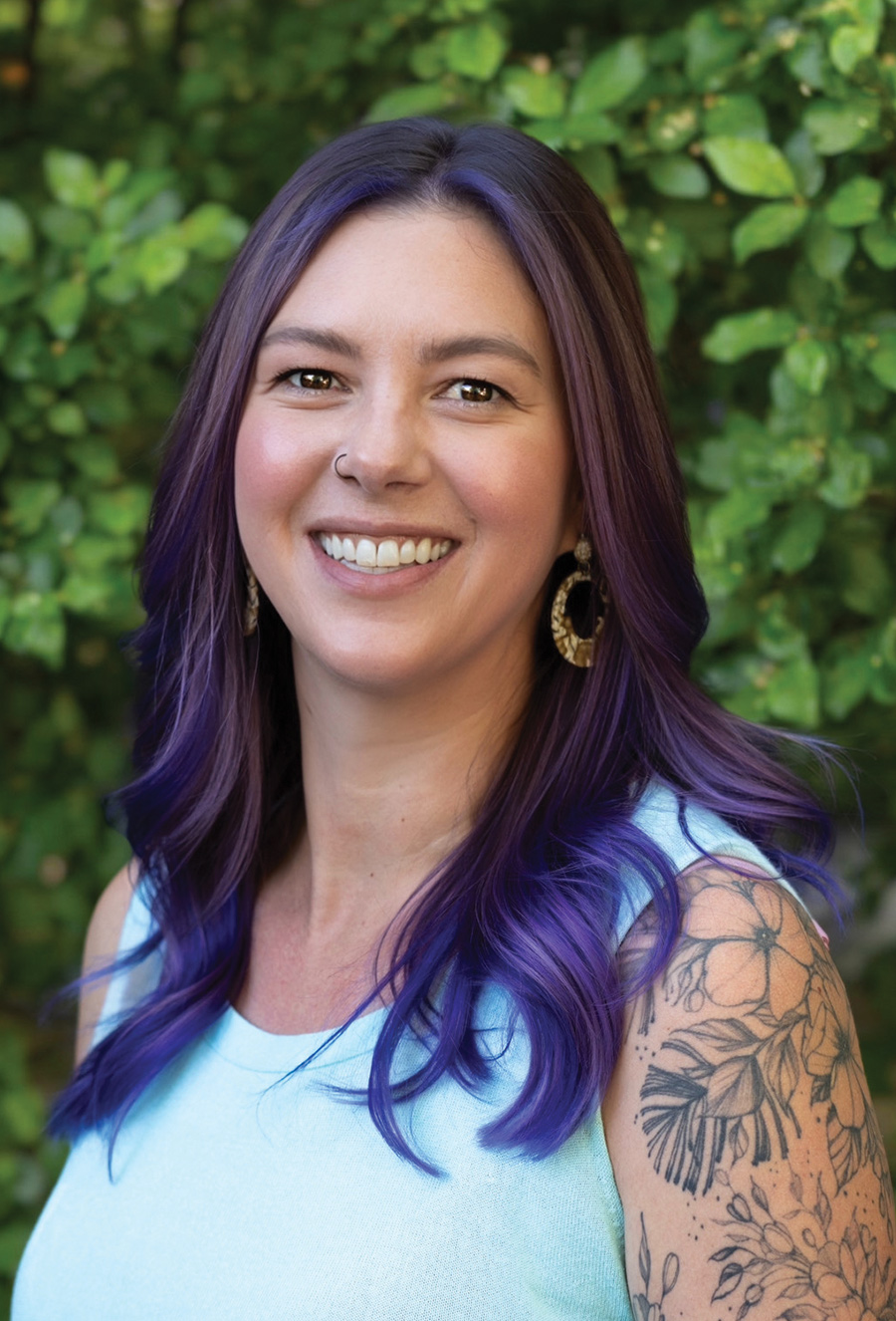 Lorna Bushong, with long purple hair, wears a light blue sleeveless top and hoop earrings as she smiles in front of a green leafy background. A floral tattoo adorns her upper arm.
