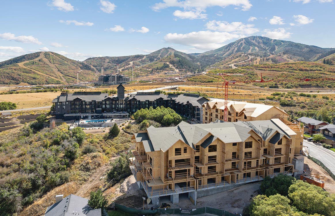 A multi-story building under construction stands in the foreground, with finished buildings, cranes, and mountains visible in the background under a partly cloudy sky.