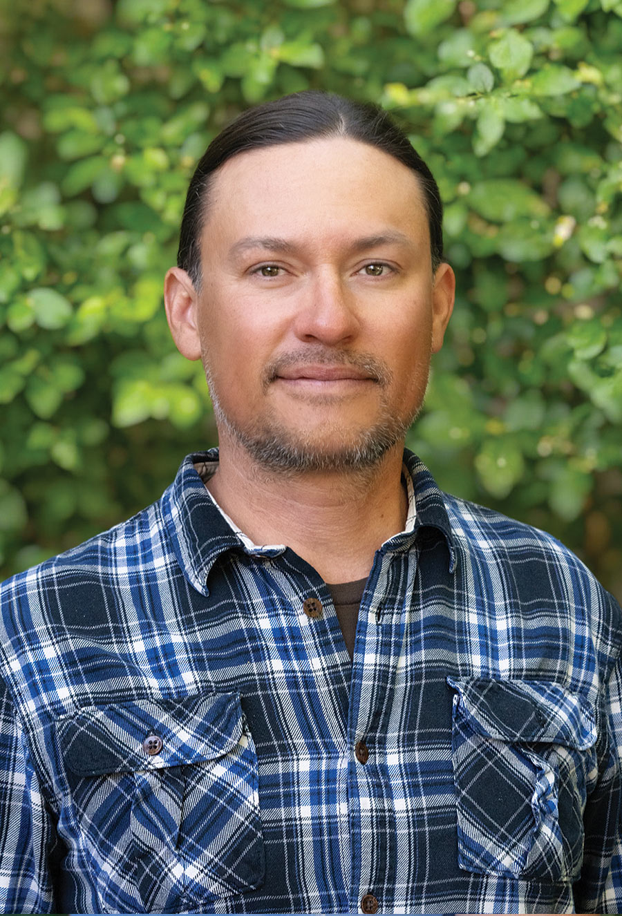 Eric Osterman, a person with long brown hair and a trimmed beard wearing a blue plaid shirt, stands in front of green foliage.