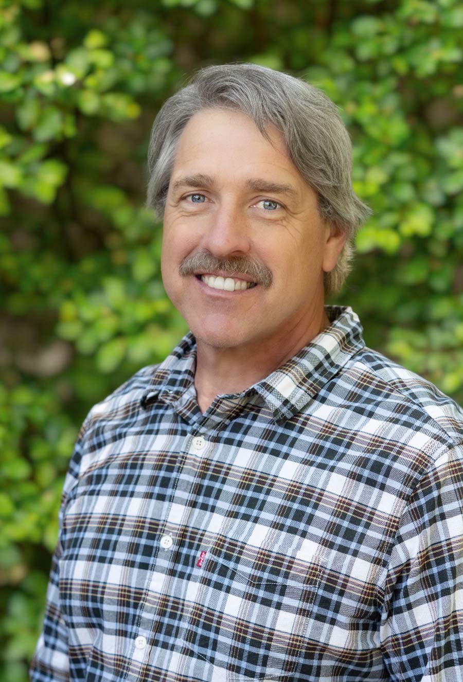 Brad Endrizal, a middle-aged man with gray hair and a mustache, stands outdoors in front of green foliage wearing a plaid shirt, smiling at the camera.