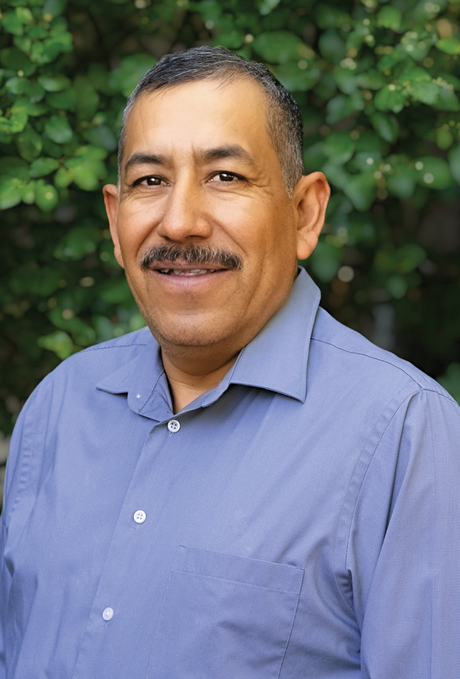 Jorge Hernandez, a man with short dark hair and a mustache, wearing a blue button-up shirt, stands in front of green leafy bushes and smiles at the camera.