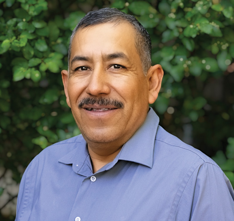 Jorge Hernandez, a man with short dark hair and a mustache, wearing a light blue collared shirt, stands in front of leafy green foliage.