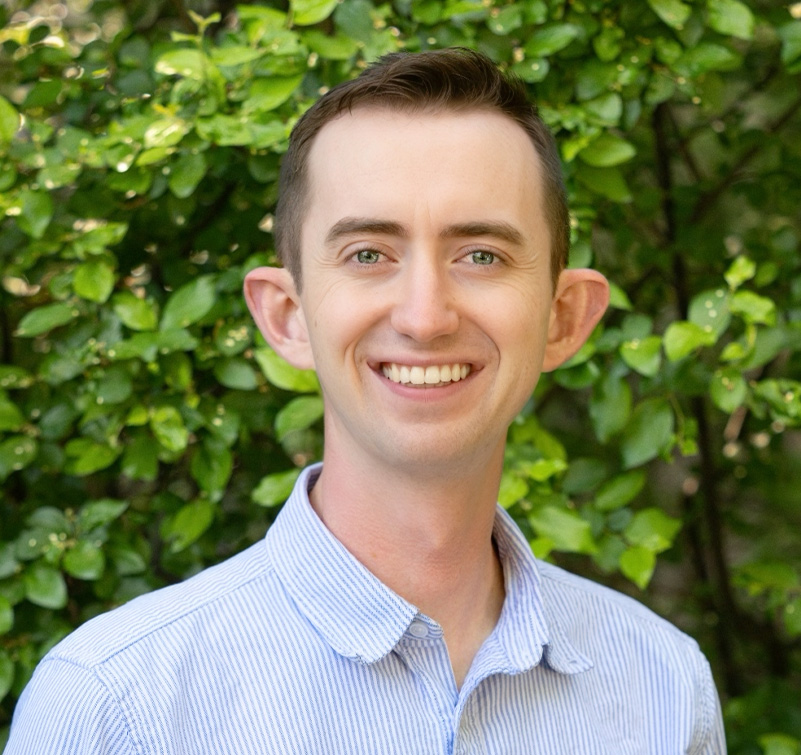 Daniel Saunders, a man with short brown hair, wearing a light blue striped shirt, smiles in front of a leafy green background.