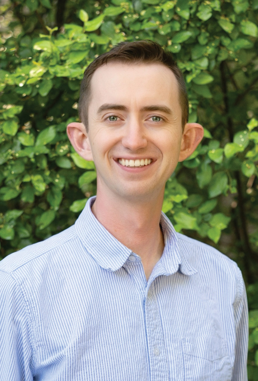 Daniel Saunders, wearing a light blue striped shirt, stands outdoors in front of green leafy bushes, smiling at the camera.