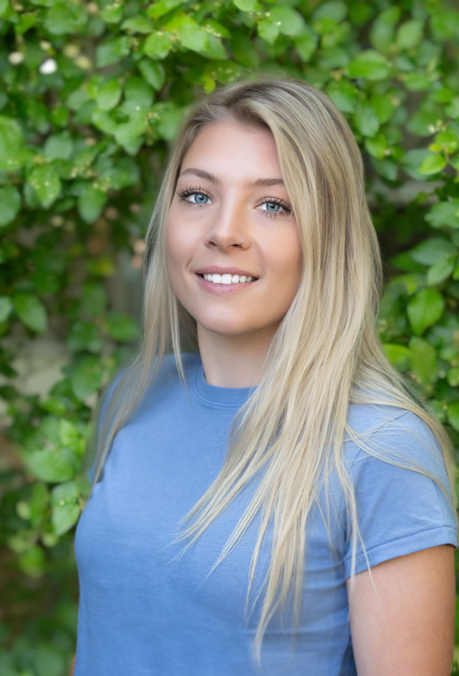 Emily McGlone, a woman with long blonde hair and a blue shirt, stands outdoors in front of leafy green foliage, looking at the camera and smiling.