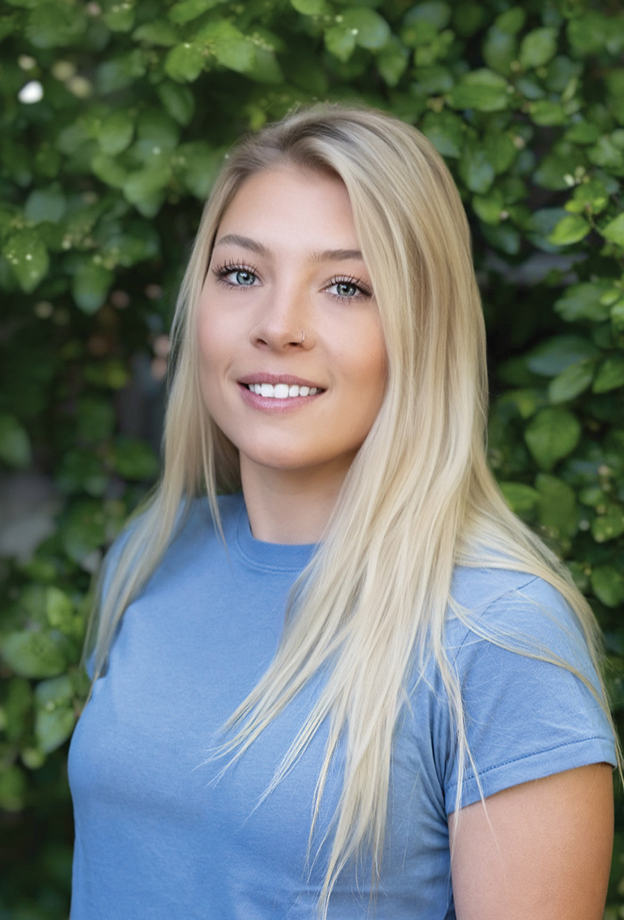 Emily McGlone, a woman with long blonde hair and a nose ring, stands outdoors in front of green foliage, smiling at the camera in her blue t-shirt.