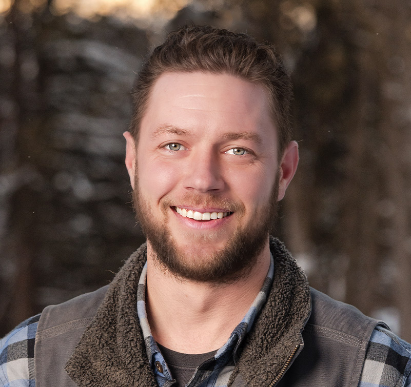 A man with a beard and short hair smiles at the camera. Austin Stoltenberg is wearing a plaid shirt and a vest, with a blurred outdoor background behind him.