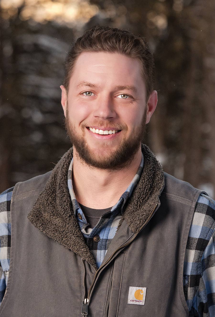 Smiling man with short brown hair and beard, Austin Stoltenberg, wearing a plaid shirt and gray vest, standing outdoors with blurred trees in the background.