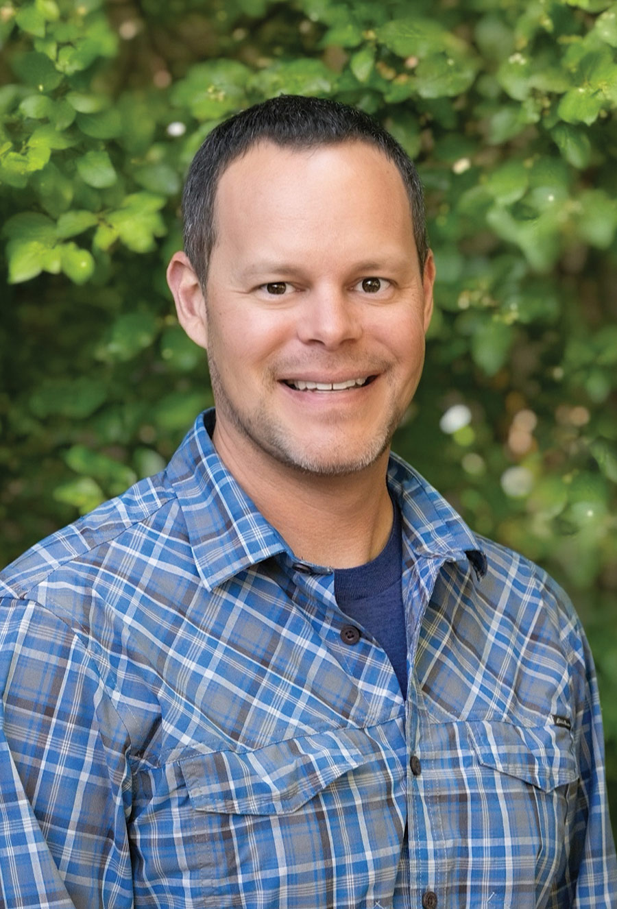 A man in a blue plaid shirt smiles outdoors in front of leafy green foliage.