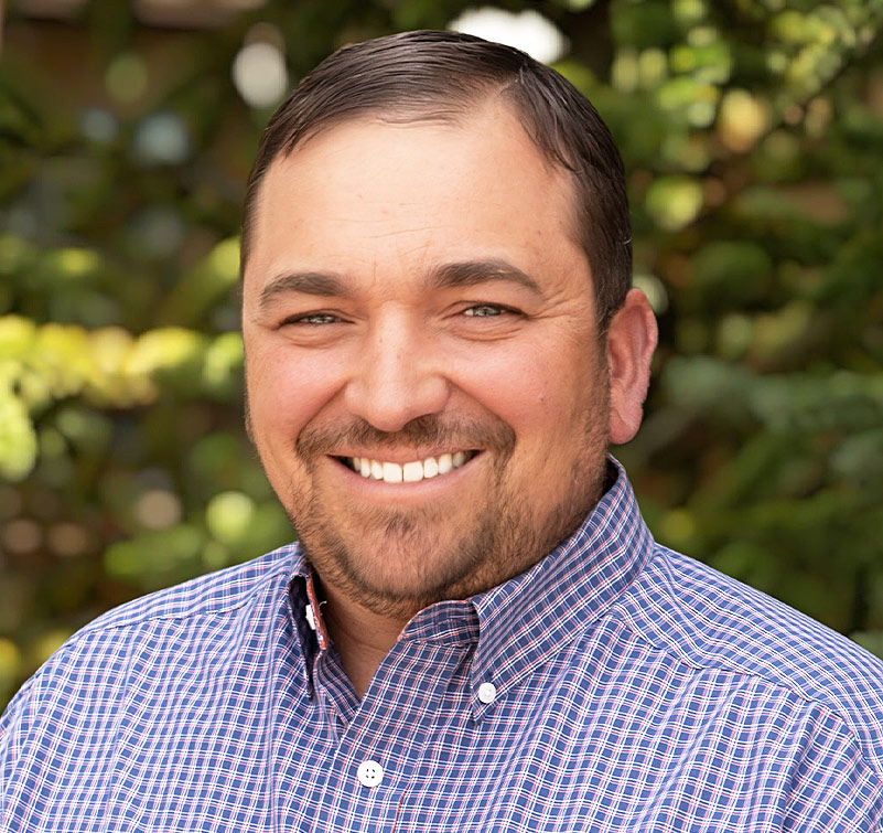 Craig Wilcox, a man with short brown hair and a trimmed beard, smiles at the camera in a plaid button-up shirt, with greenery in the background.