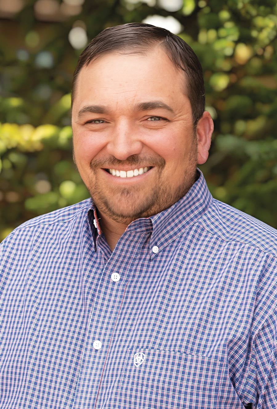 Craig Wilcox, a man with short brown hair and a beard, smiles at the camera in a checked button-up shirt. Green foliage is blurred in the background.
