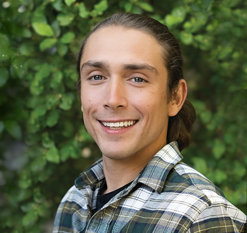 A young man with long brown hair in a ponytail, wearing a plaid shirt, smiles in front of green foliage.
