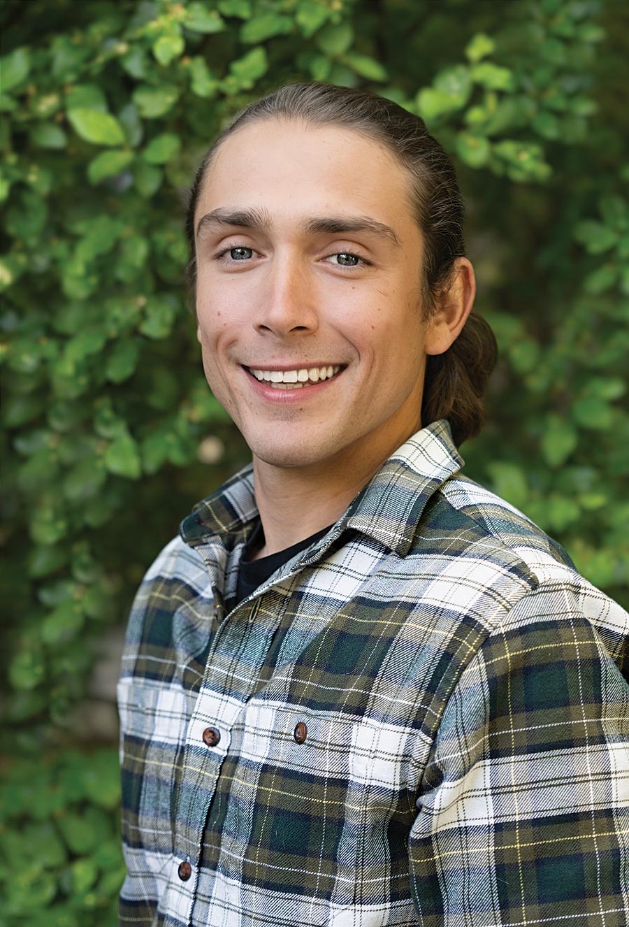 A person in a green plaid shirt stands outdoors in front of leafy greenery, smiling at the camera.
