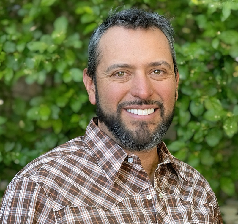 Alexander Trujillo, a man with short dark hair and a beard, wearing a brown plaid shirt, smiles outdoors with green foliage in the background.
