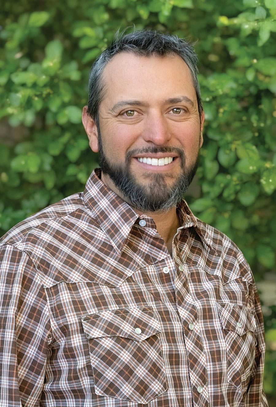 Alexander Trujillo, a man with short dark hair and a beard, wearing a brown plaid shirt, stands outdoors in front of green foliage, smiling at the camera.