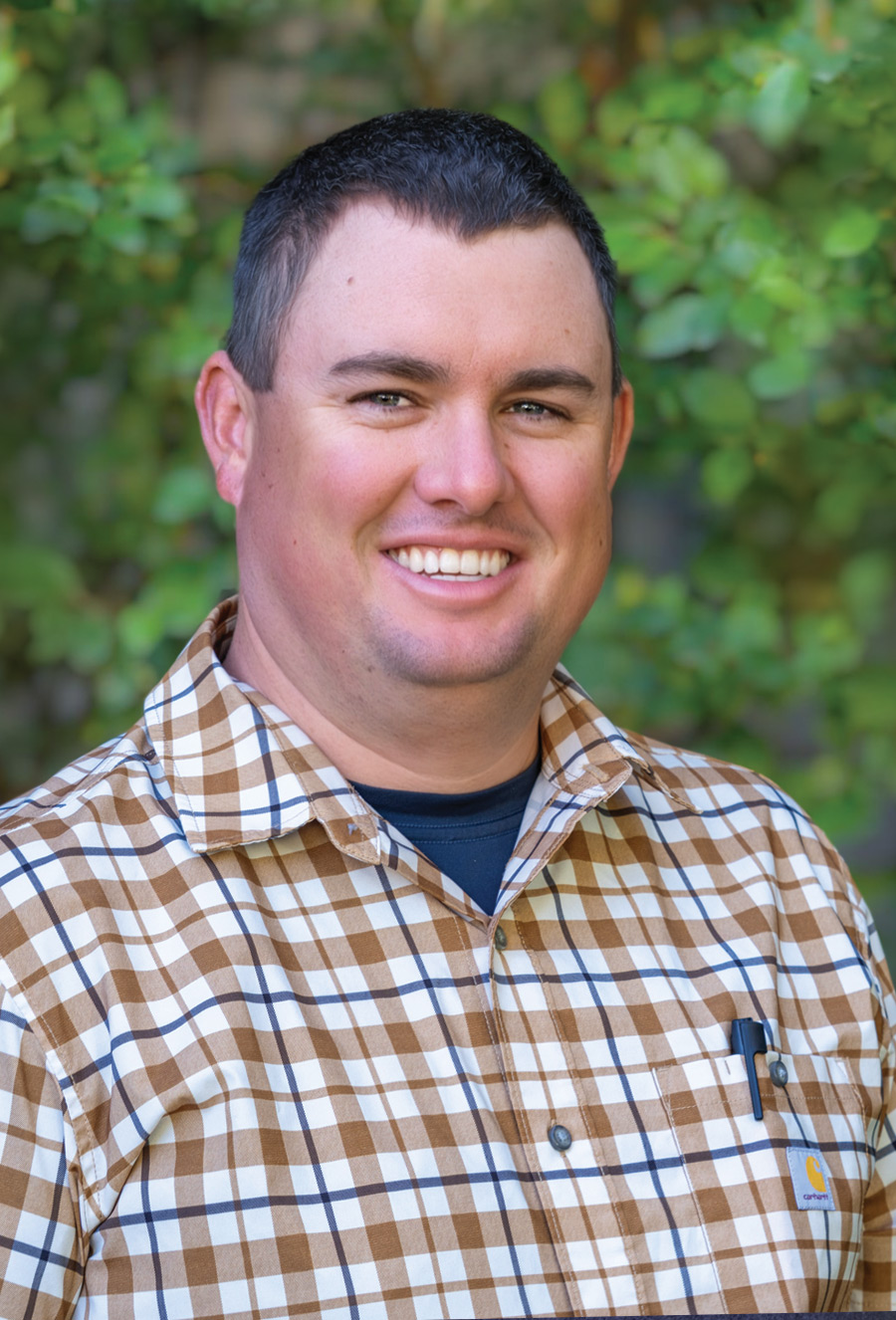 Brendan Curnow, a man with short dark hair wearing a brown and white plaid shirt, smiles at the camera while standing outdoors with green foliage in the background.