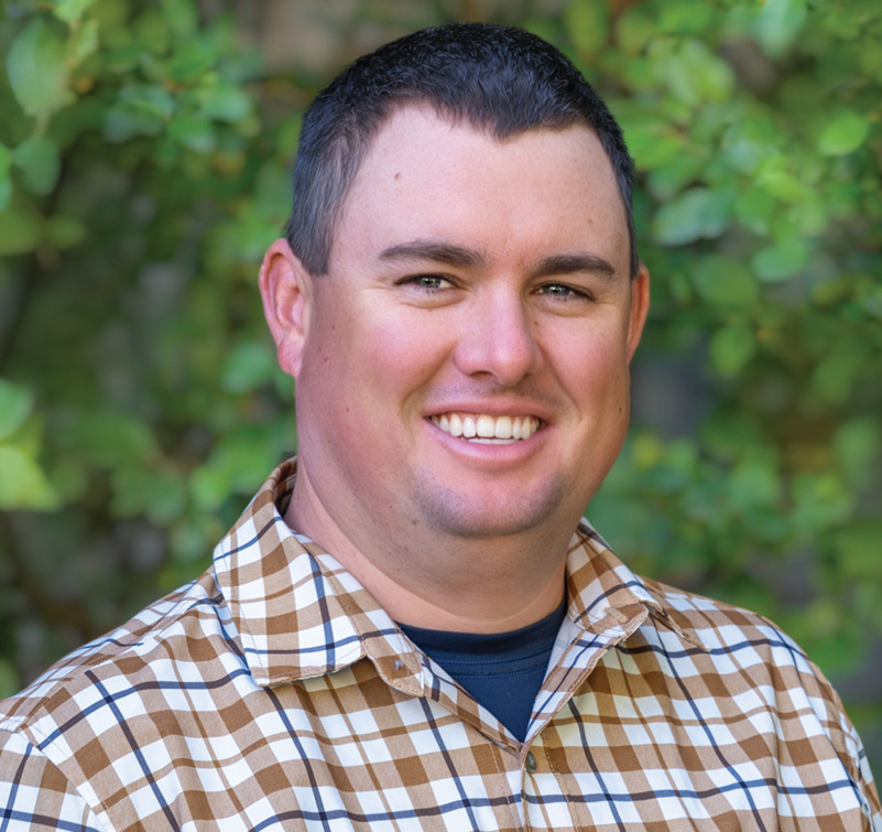 Brendan Curnow, a man with short dark hair, wearing a brown and white plaid shirt, smiles in front of green foliage.