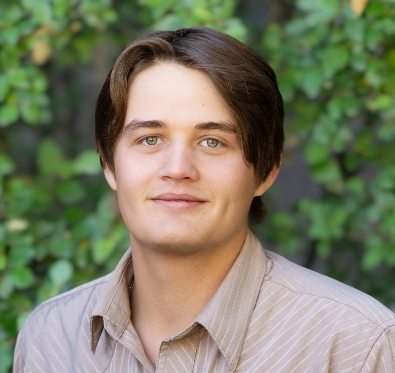 Andrew Dowdy, a young man with light skin and short brown hair, wears a beige striped shirt and stands in front of a leafy green background.