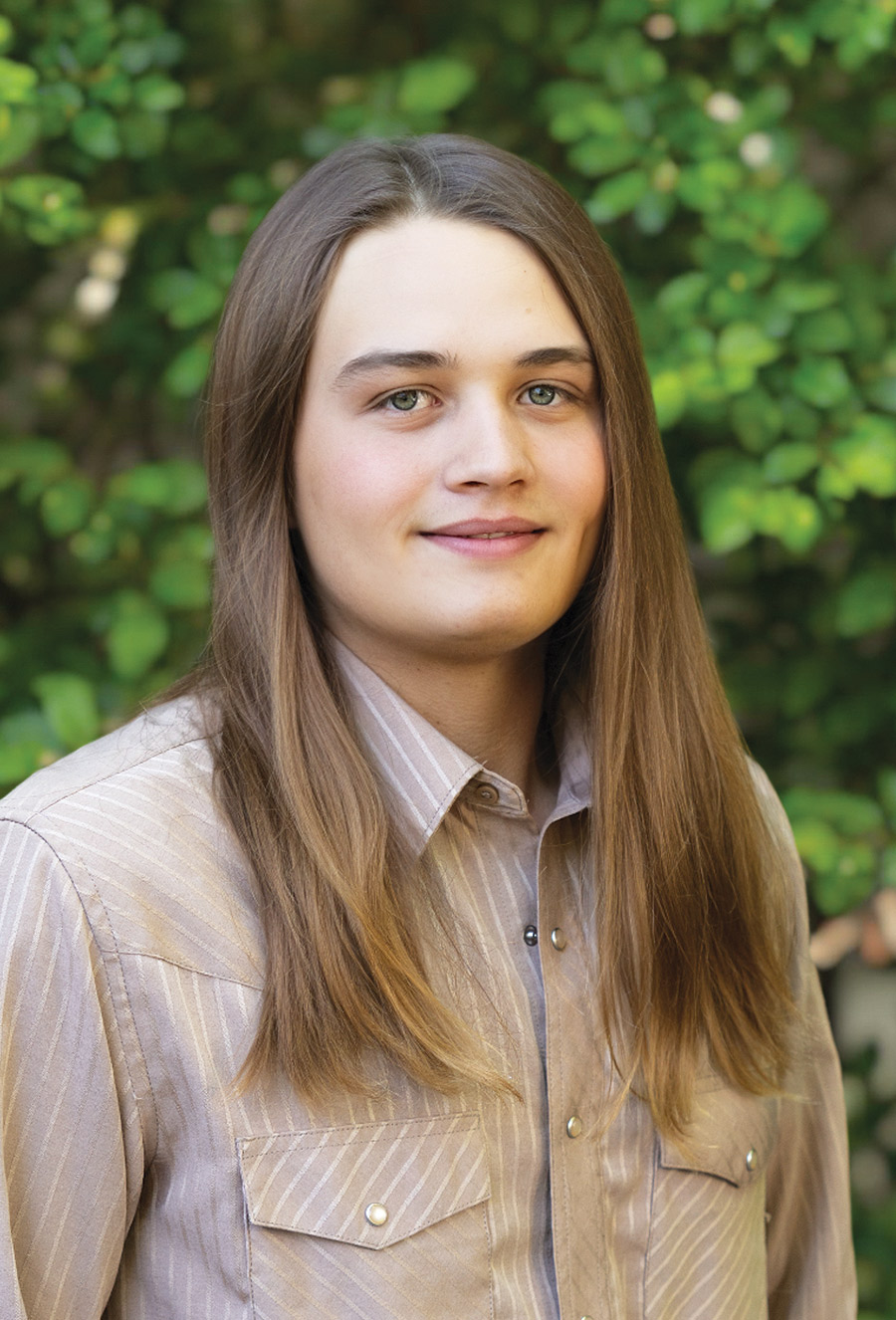 A young person with long brown hair and a beige button-up shirt stands in front of green foliage, looking at the camera—an image that could easily feature Andrew Dowdy.