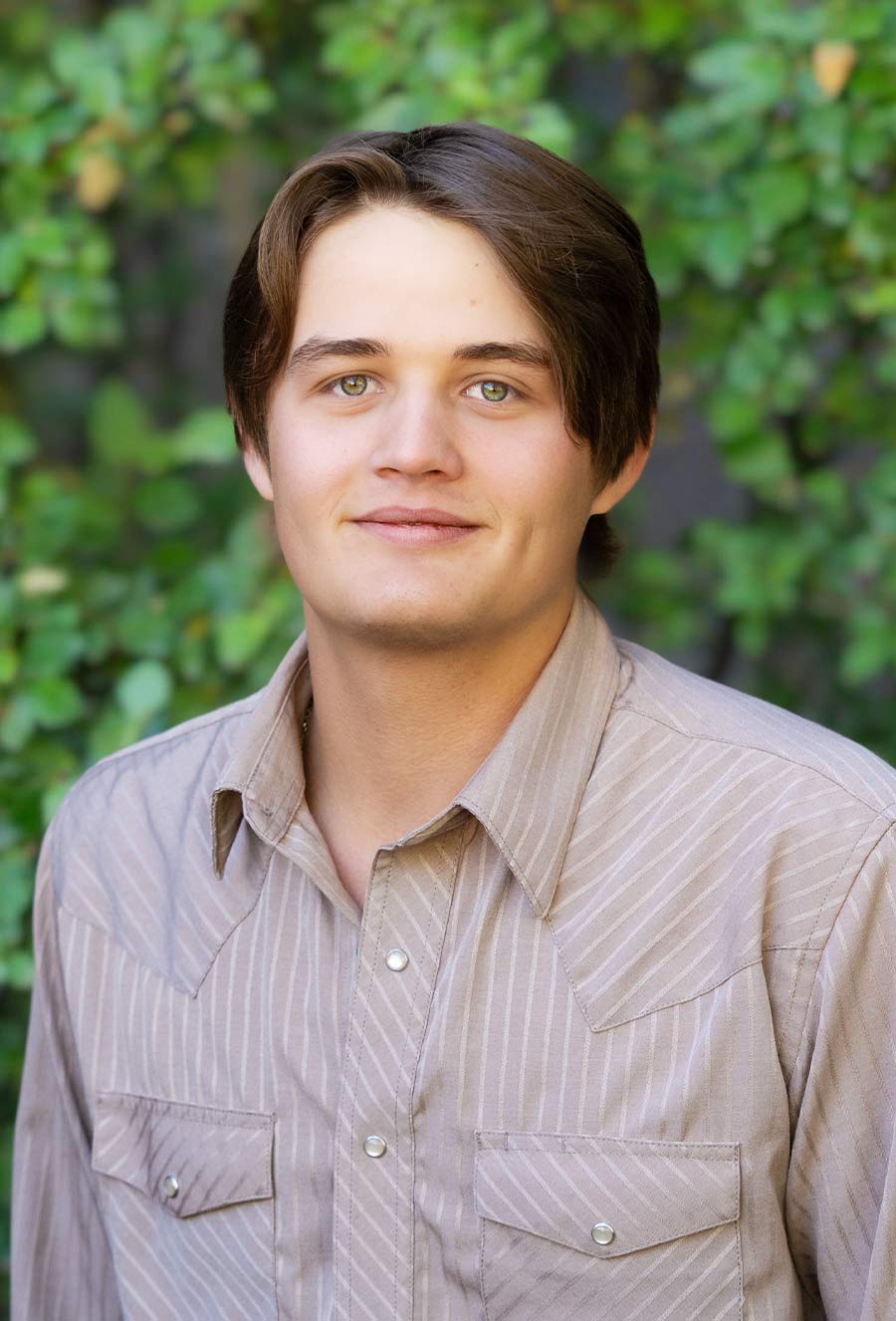 Young man with light skin, brown hair, and green eyes wearing a tan striped button-up shirt, standing in front of a leafy green background—Andrew Dowdy.