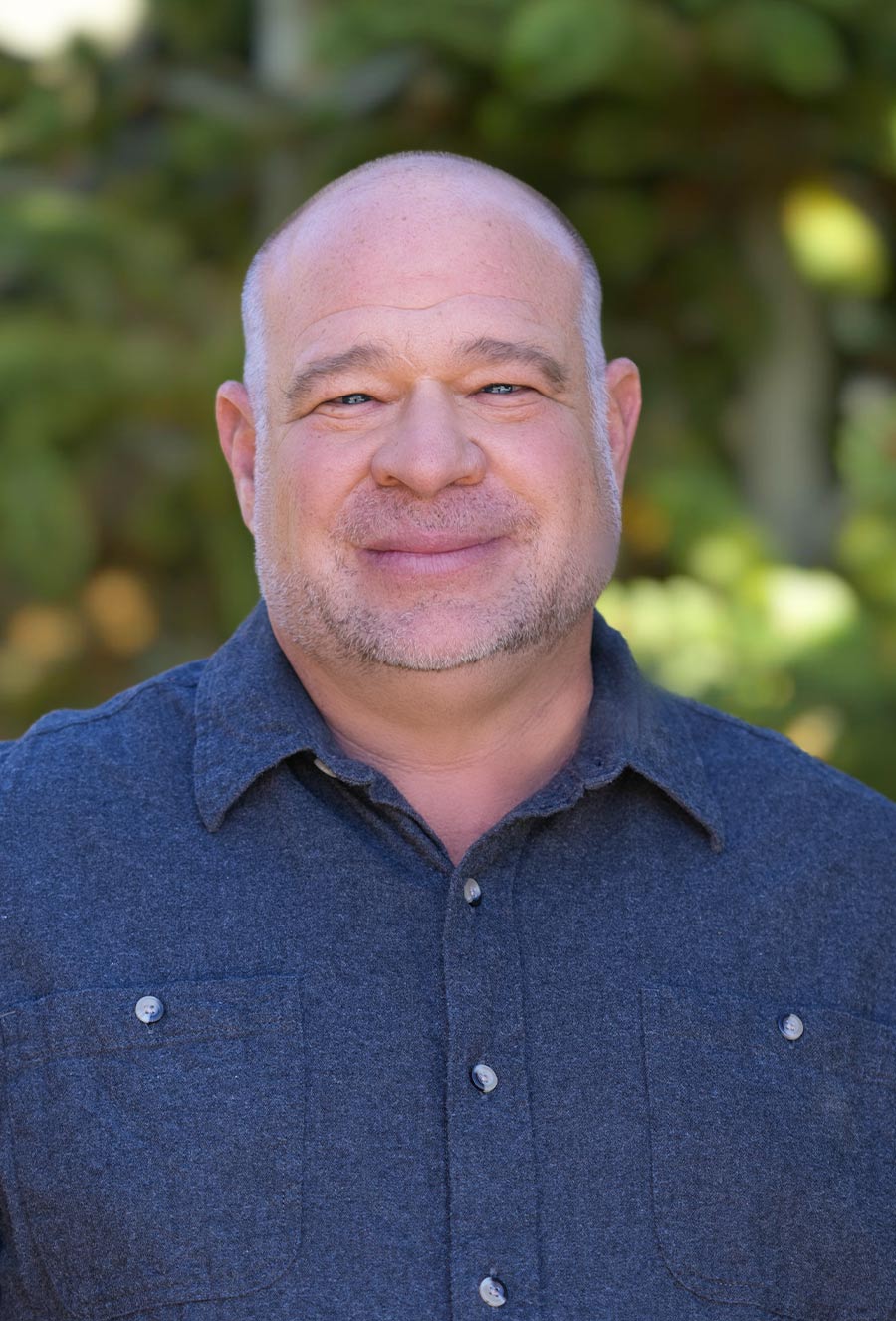 A middle-aged man with a shaved head and short beard, the Blair Hill Construction Superintendent, stands outdoors in a blue button-up shirt with greenery in the background.