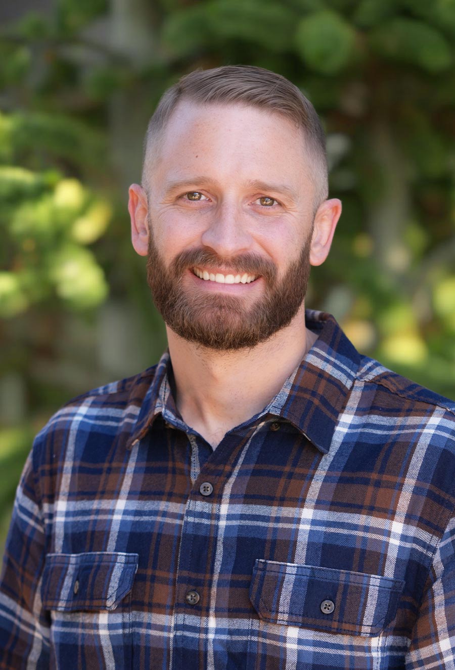 Taylor Remington, a man with short hair and a beard, stands outdoors in a blue and brown plaid shirt, smiling at the camera with greenery in the background.