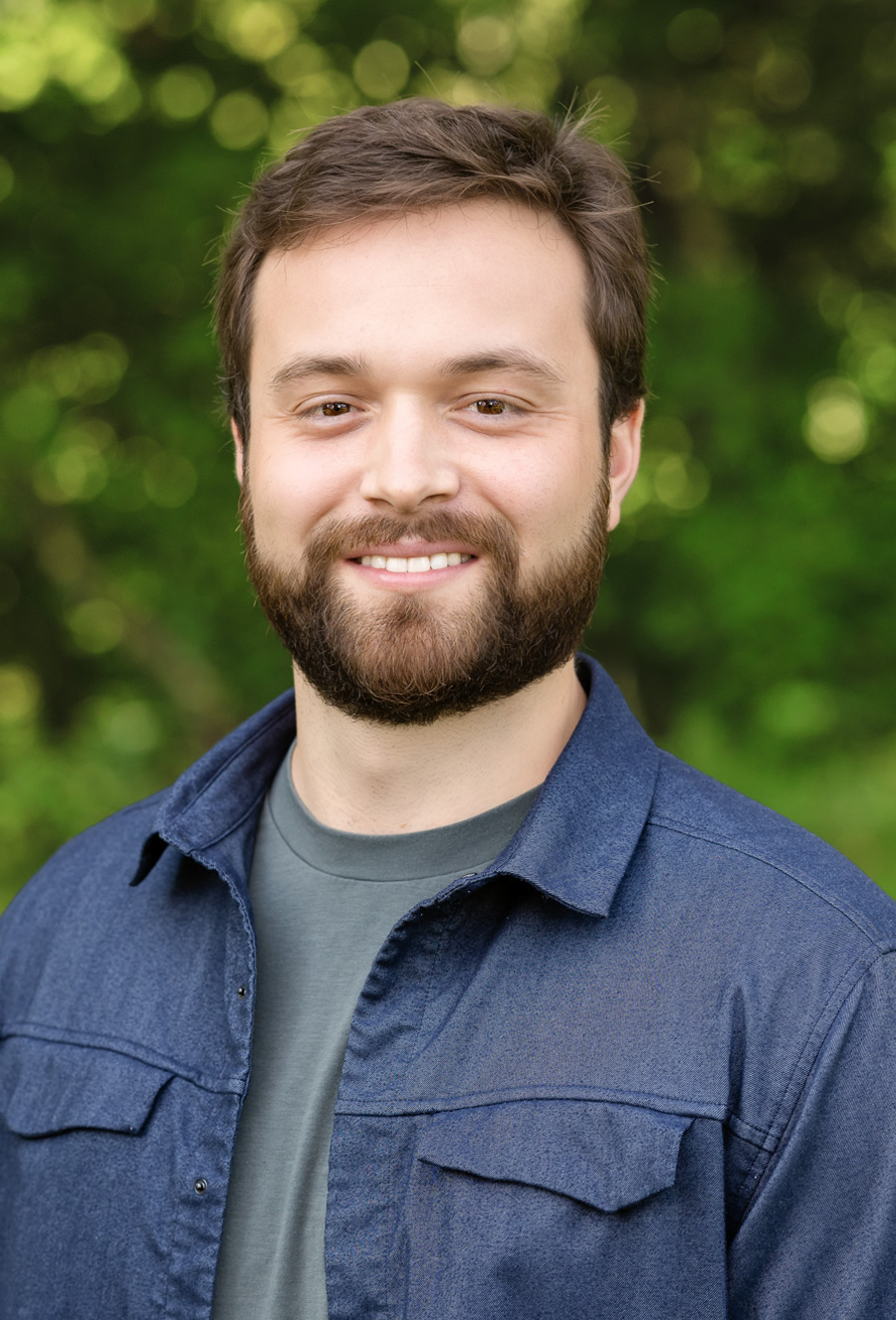 A man with short brown hair and a beard is smiling, wearing a dark blue shirt over a gray t-shirt, standing outdoors with green foliage in the background.