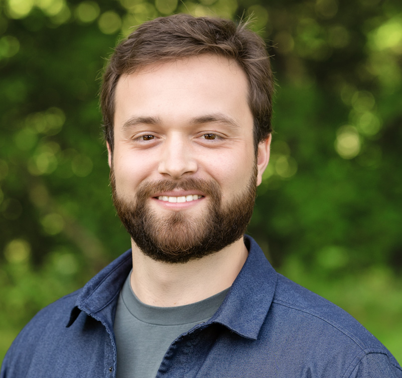 A man with a beard and brown hair smiles at the camera while standing outdoors with green foliage in the background.
