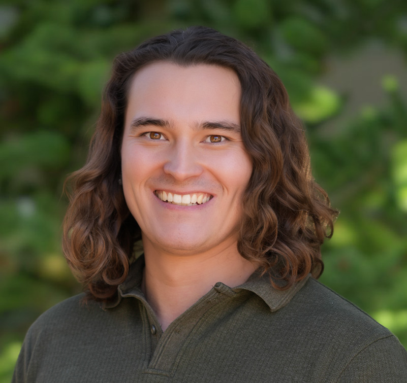 A person with long brown hair wearing a green collared shirt smiles in front of a blurred leafy background.