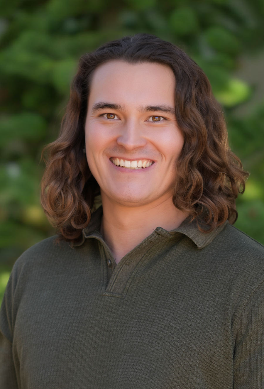 A person with long, wavy brown hair and a green collared shirt smiles at the camera with a blurred green background.