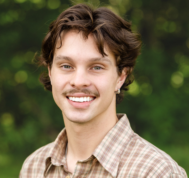 A young man with wavy brown hair, a mustache, and an earring smiles at the camera while standing outdoors in front of a blurred green background—Nolan Blessington exudes effortless charm.
