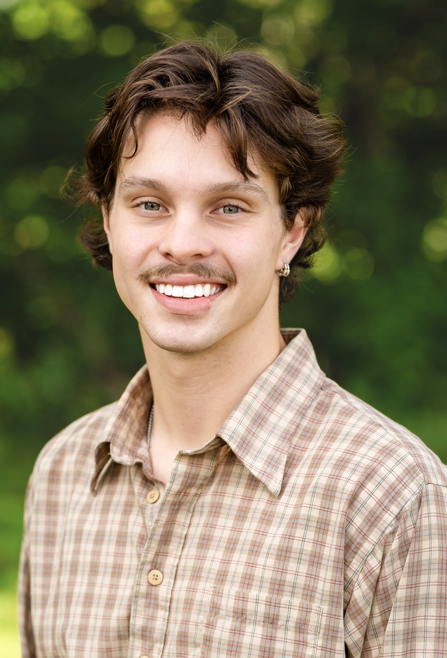 Nolan Blessington, a young man with wavy brown hair and a mustache, wears a plaid button-up shirt while smiling outdoors against a blurred green background.