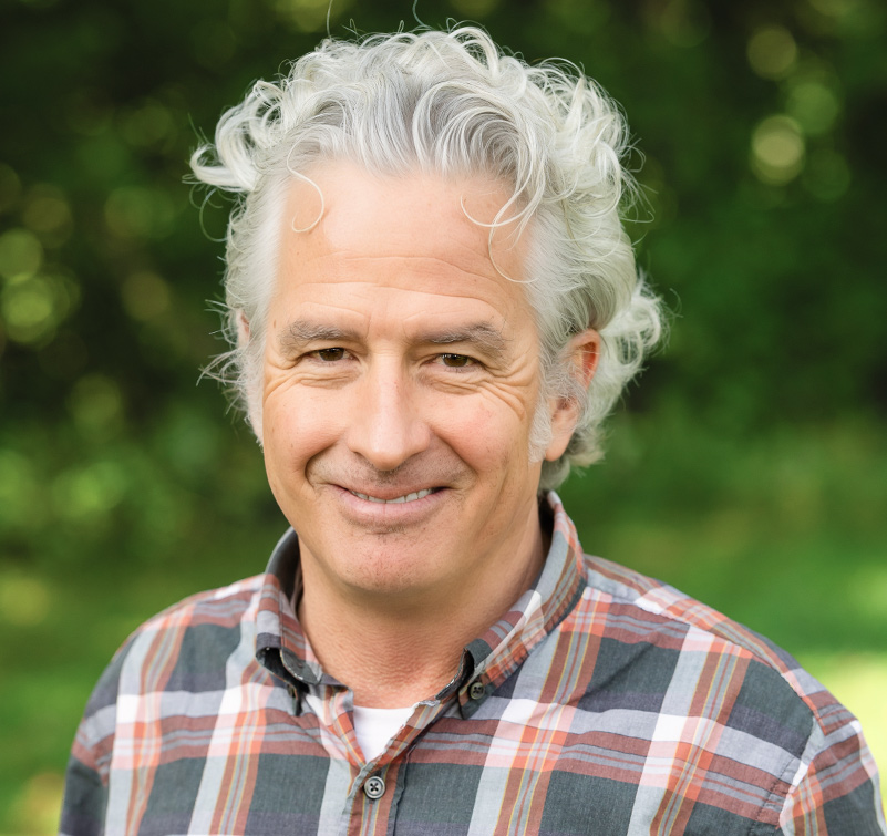 A middle-aged man with curly gray hair smiles outdoors, wearing a plaid shirt. Green foliage is blurred in the background.