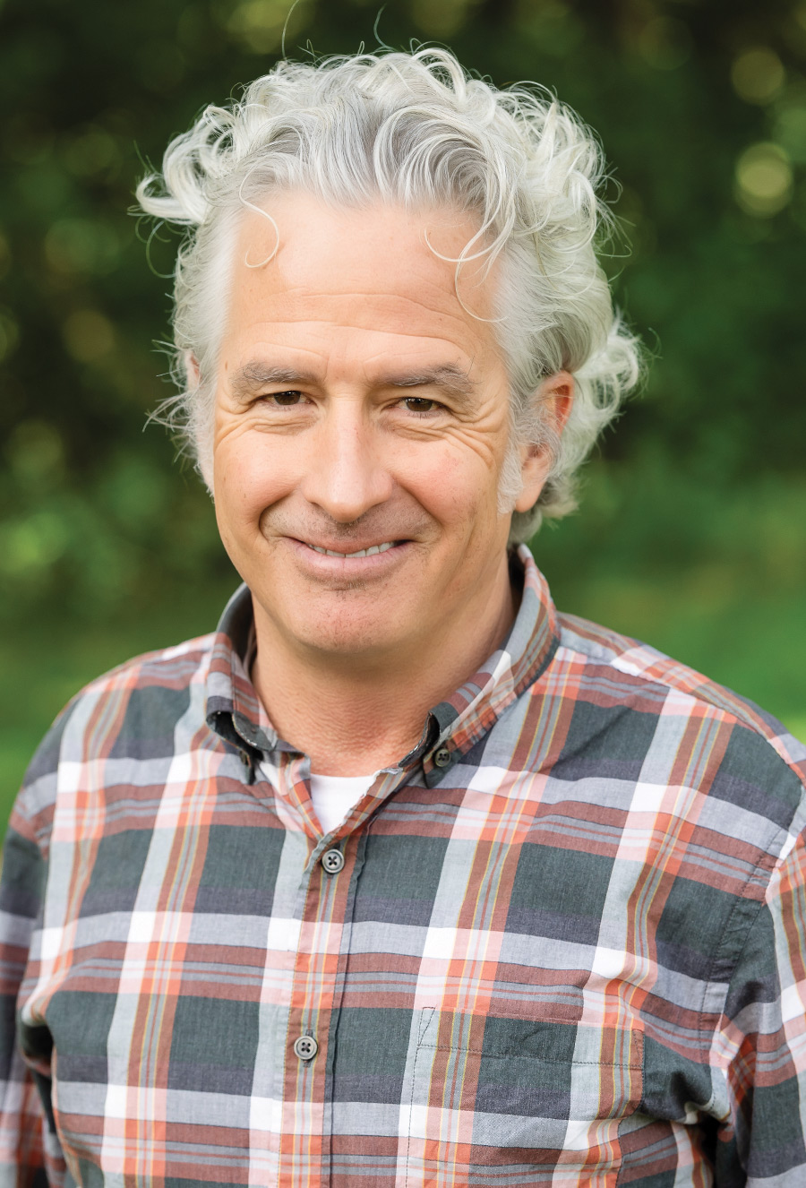 An older man with gray, curly hair wearing a plaid shirt stands outdoors, smiling at the camera with a blurred green background.