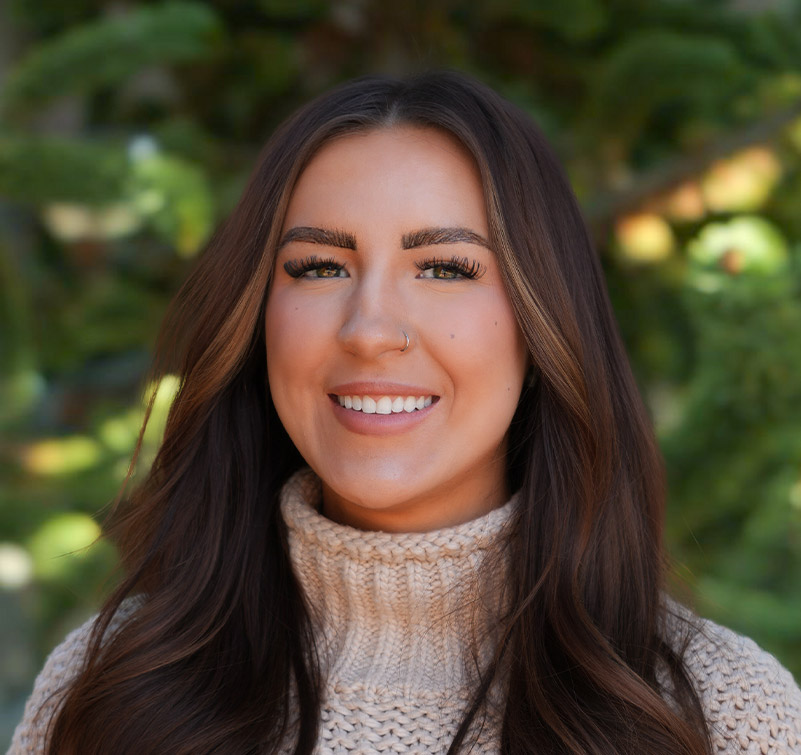 Woman with long brown hair wearing a beige knit sweater, smiling outdoors with green foliage in the background.
