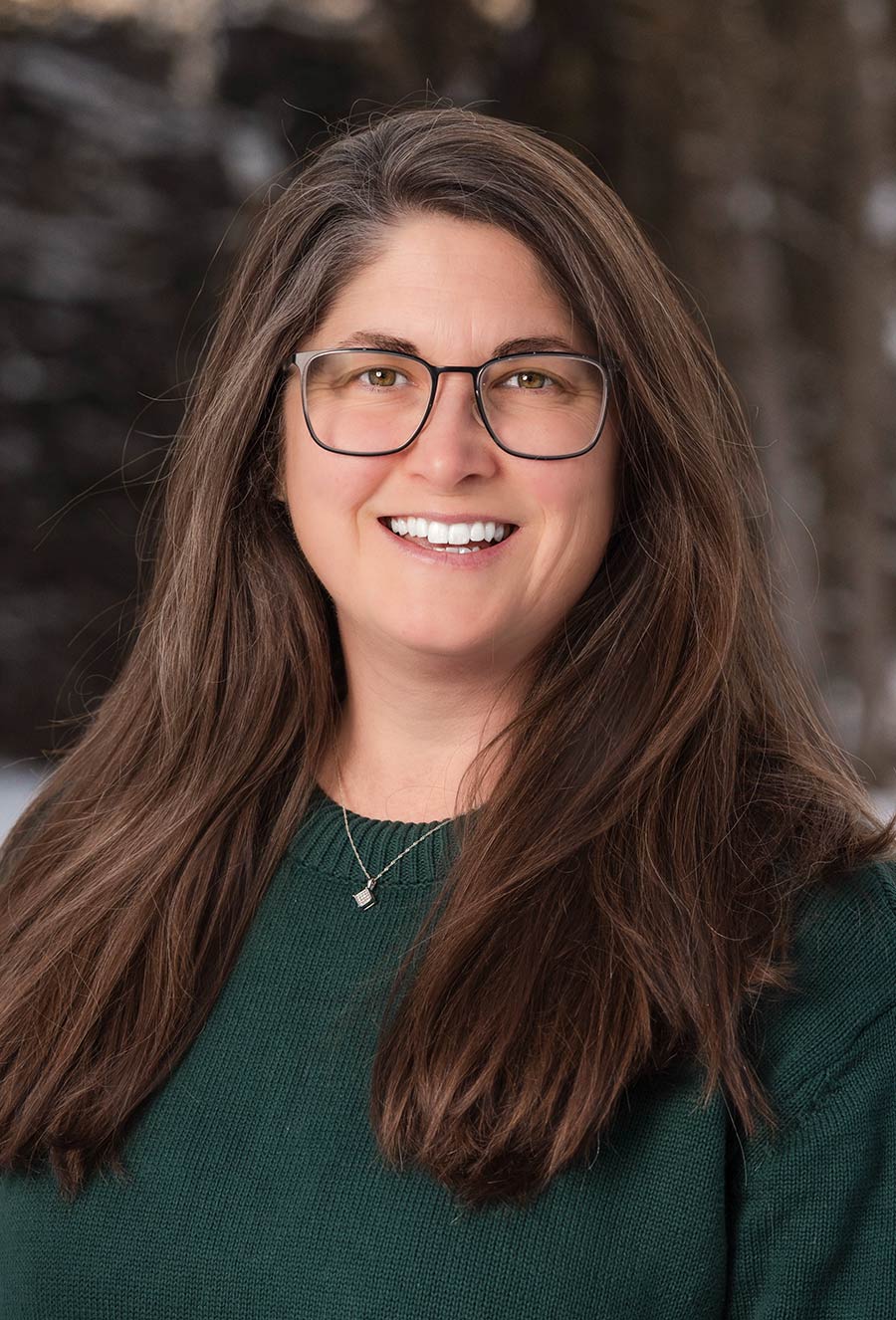 Woman with long brown hair and glasses wearing a green sweater, smiling outdoors with a blurred background.