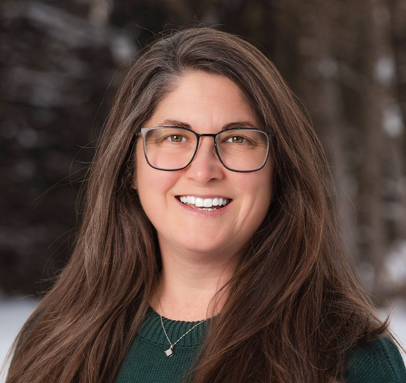 Woman with long brown hair and glasses, wearing a green sweater and a necklace, smiling outdoors with a blurred winter background.