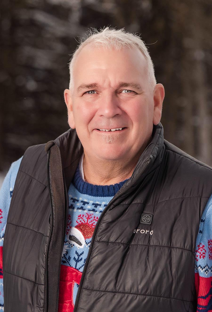 Eddie McEvoy, a middle-aged man with short gray hair, smiles at the camera, wearing a colorful sweater and a black puffy vest with a blurred outdoor background.