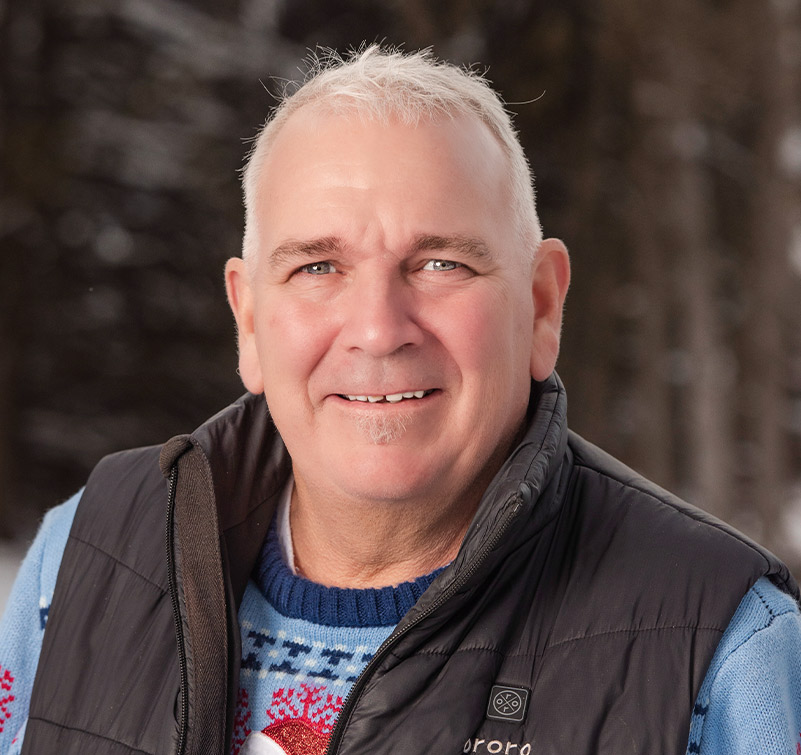 Eddie McEvoy, an older man with short white hair, smiles outdoors in a blue patterned sweater and black puffer vest. Snow and blurred trees create a wintry backdrop.