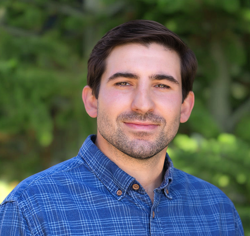 Man with dark hair and beard, wearing a blue plaid shirt, stands outdoors in front of blurred green foliage, looking at the camera with a neutral expression.