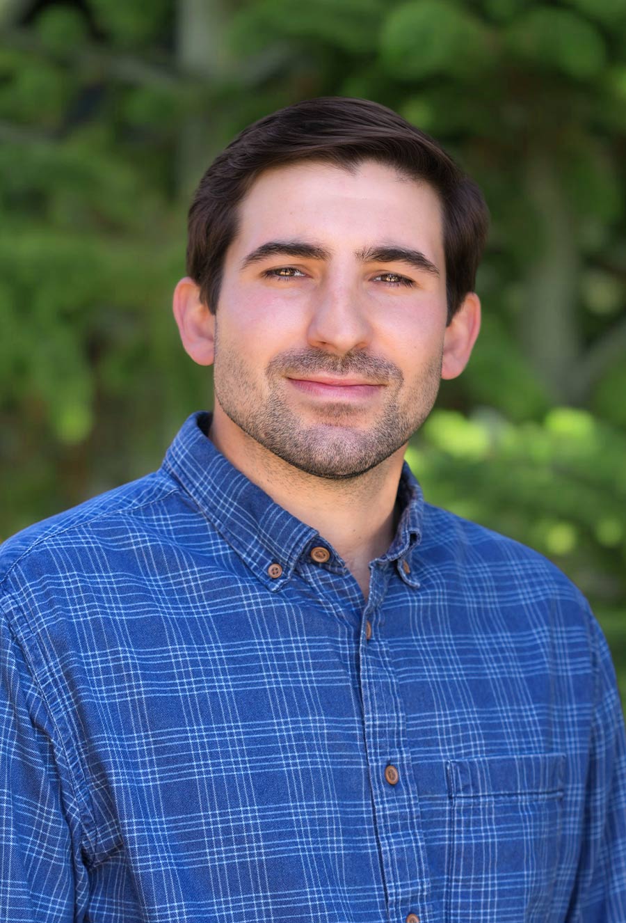 A man with short dark hair and a trimmed beard wearing a blue plaid shirt stands outdoors in front of green foliage.