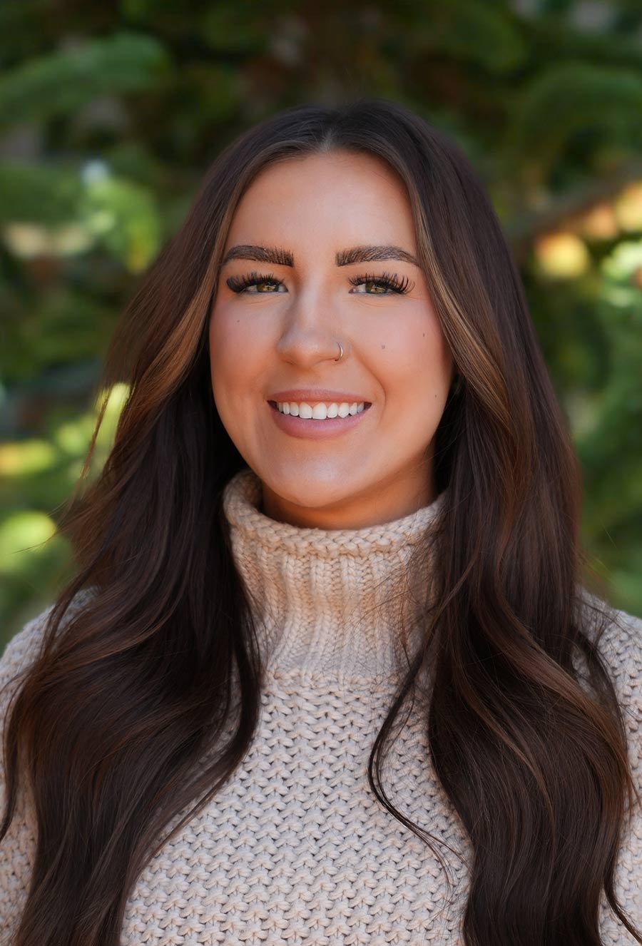 A woman with long brown hair, wearing a beige knit turtleneck sweater, smiles outdoors with greenery in the background.