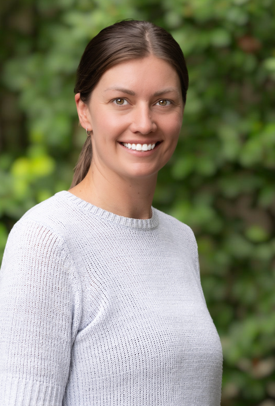 Marion Thurston, a woman with brown hair in a ponytail, wearing a light gray sweater, stands outdoors in front of a blurred green leafy background, smiling at the camera.
