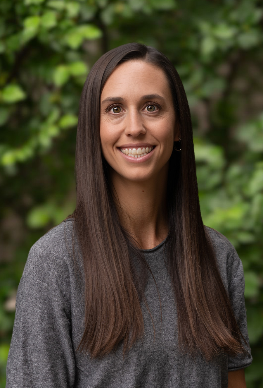 Woman with straight brown hair and a gray shirt smiles at the camera in front of a blurred green leafy background.