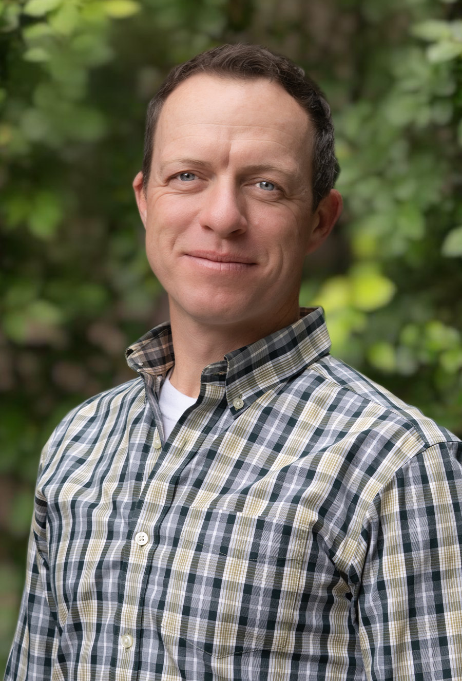 A man with short brown hair wearing a plaid shirt stands outdoors in front of a blurred background of green foliage.