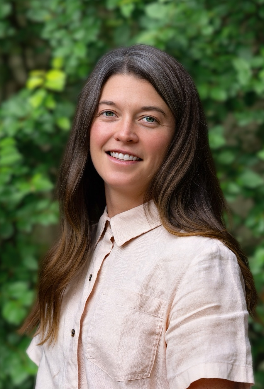 A woman with long brown hair and a beige button-up shirt stands outdoors in front of green foliage, smiling at the camera.