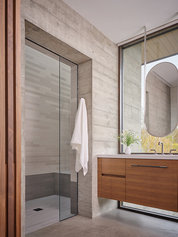 Modern bathroom with a glass-enclosed shower, light gray tiled walls, floating wood vanity, oval mirror, and a white towel hanging on a hook. Large window lets in natural light.
