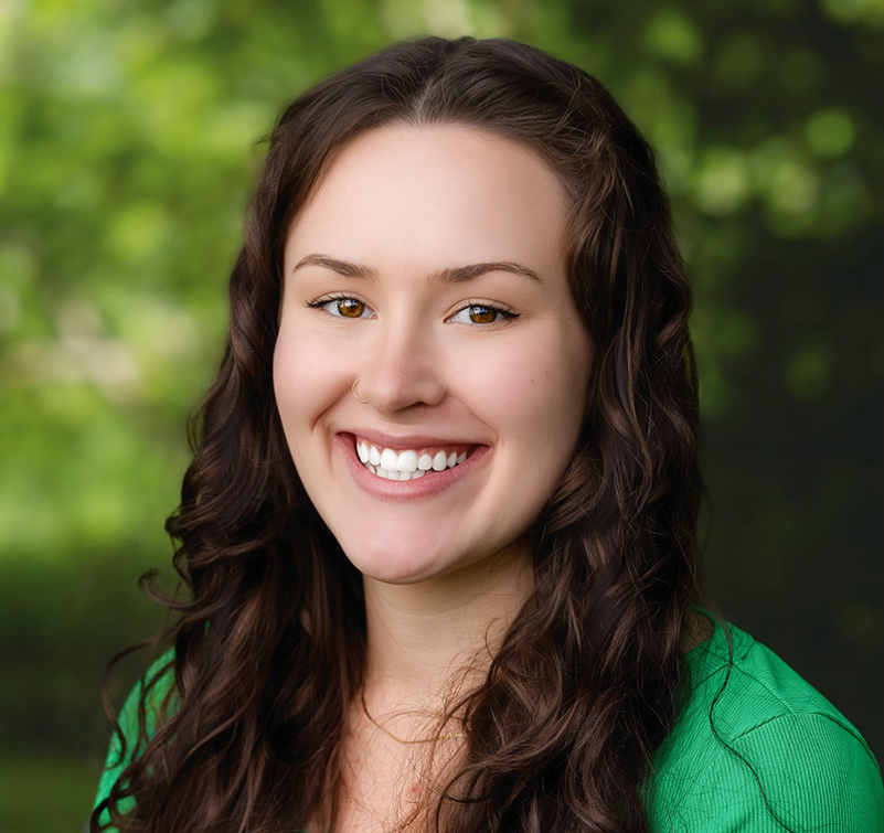 A woman with long curly brown hair and a nose ring smiles at the camera, wearing a green shirt. Reminiscent of Abigail Williams, the background is blurred greenery.